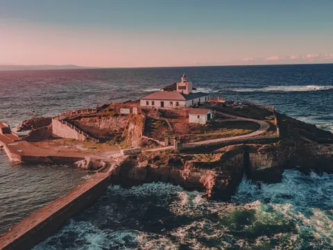 Vista aérea del faro de Tapia de Casariego al atardecer, rodeado por el mar Cantábrico y los acantilados de la costa asturiana.
