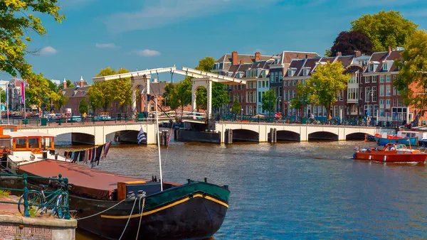 Vista de un canal en Ámsterdam con barcos amarrados y gente navegando, al fondo el puente levadizo blanco Magere Brug y casas típicas holandesas rodeadas de árboles.