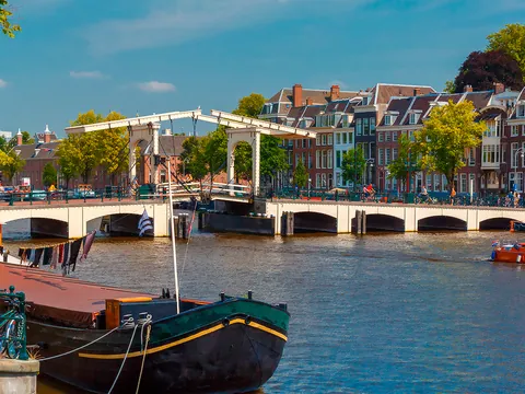 Vista de un canal en Ámsterdam con barcos amarrados y gente navegando, al fondo el puente levadizo blanco Magere Brug y casas típicas holandesas rodeadas de árboles.