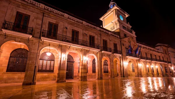 Ayuntamiento de Oviedo iluminado de noche con reflejos en el suelo mojado.