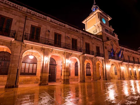 Ayuntamiento de Oviedo iluminado de noche con reflejos en el suelo mojado.