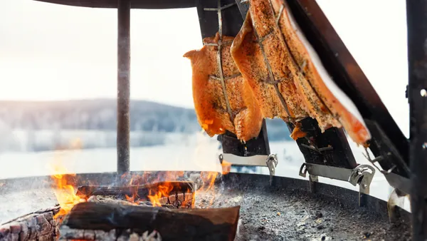 Filetes de salmón sazonados asándose al fuego en una parrilla tradicional al aire libre, con brasas encendidas y paisaje invernal de fondo.