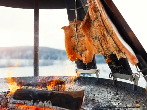 Filetes de salmón sazonados asándose al fuego en una parrilla tradicional al aire libre, con brasas encendidas y paisaje invernal de fondo.