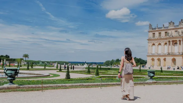 Una mujer con un vestido blanco camina hacia el Palacio de Versalles, mostrando su majestuosa fachada exterior.