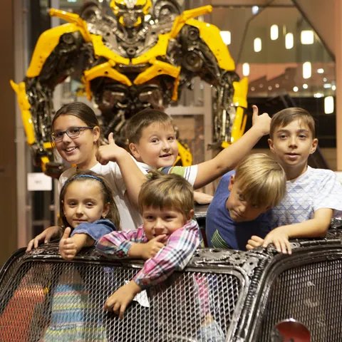 Niños dentro de un coche hecho de hierro con una figura de Bumblebee detras en la Galería de las Figuras de Hierro de Praga