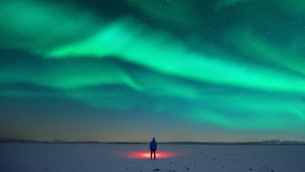 Persona de pie sobre nieve iluminada por luz roja, observando auroras boreales verdes que cubren el cielo estrellado.
