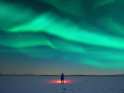 Persona de pie sobre nieve iluminada por luz roja, observando auroras boreales verdes que cubren el cielo estrellado.