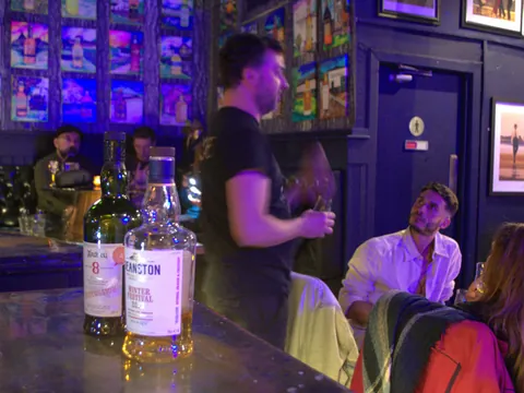 Sesión de cata de whisky escocés en un bar, con dos botellas de whisky en primer plano y personas participando en la actividad.
