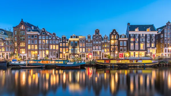 Casas tradicionales neerlandesas iluminadas al anochecer junto a un canal de Ámsterdam, con barcos estacionados y reflejos de los edificios en el agua.
