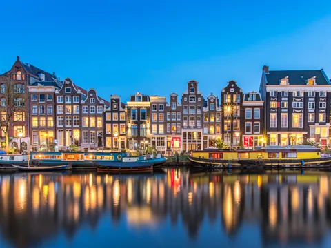Casas tradicionales neerlandesas iluminadas al anochecer junto a un canal de Ámsterdam, con barcos estacionados y reflejos de los edificios en el agua.