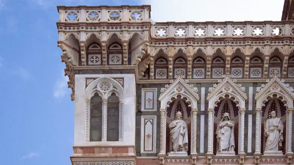 Detalle arquitectónico de la fachada del Duomo de Florencia, con ventanas góticas, esculturas religiosas en nichos y decoración ornamental en mármol blanco, verde y rosa.