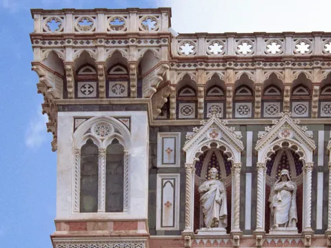 Detalle arquitectónico de la fachada del Duomo de Florencia, con ventanas góticas, esculturas religiosas en nichos y decoración ornamental en mármol blanco, verde y rosa.