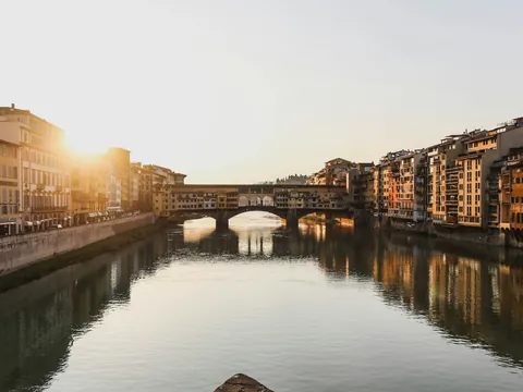 Vista del Ponte Vecchio en Florencia al atardecer, con el sol iluminando los edificios históricos a ambos lados del río Arno y reflejos en el agua.