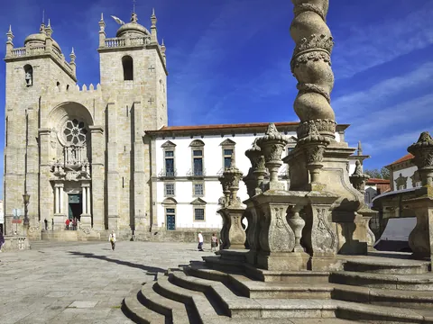 Fachada de la Catedral de Oporto y su plaza con columna barroca bajo un cielo azul.