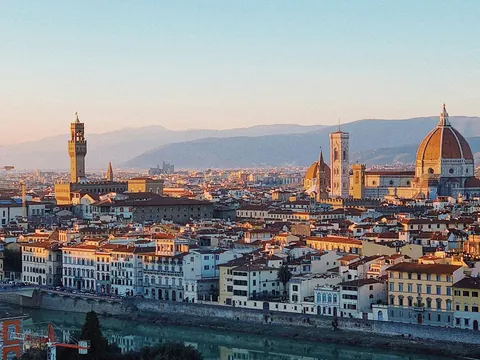 Vista panorámica de Florencia, Italia, con el río Arno en primer plano, la Catedral de Santa María del Fiore con su cúpula icónica, y la torre del Palazzo Vecchio al fondo, rodeados de edificios históricos y montañas.