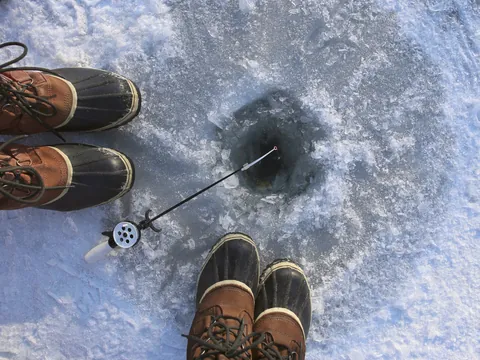 Dos pares de botas sobre hielo junto a un agujero para pesca en un lago congelado, con caña de pescar apoyada en el borde, en un paisaje invernal.