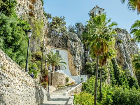 Escalinata y vegetación que llevan al castillo y campanario de Guadalest entre montañas.