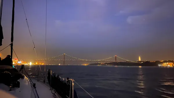 Vista nocturna del Puente 25 de Abril en Lisboa iluminado, con reflejos de luces en el río Tajo bajo un cielo oscuro.