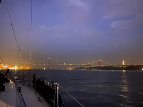 Vista nocturna del Puente 25 de Abril en Lisboa iluminado, con reflejos de luces en el río Tajo bajo un cielo oscuro.