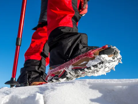 Primer plano de una bota con crampones sobre nieve, junto a un bastón de senderismo, con cielo azul despejado al fondo.