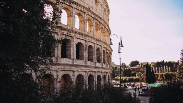 Vista lateral del Coliseo de Roma entre vegetación, con el área arqueológica al fondo.