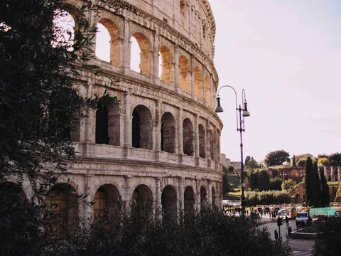Vista lateral del Coliseo de Roma entre vegetación, con el área arqueológica al fondo.