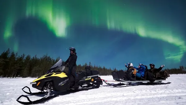 Motonieve sobre nieve con trineo y personas, bajo auroras boreales verdes en el cielo nocturno.