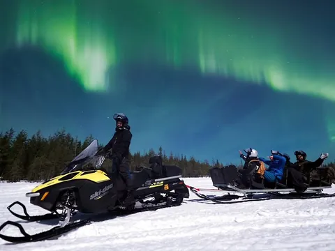 Motonieve sobre nieve con trineo y personas, bajo auroras boreales verdes en el cielo nocturno.