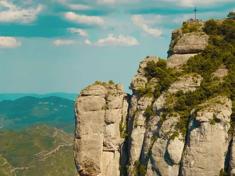 Peñasco de Montserrat con cruz en la cima y montañas verdes al fondo.