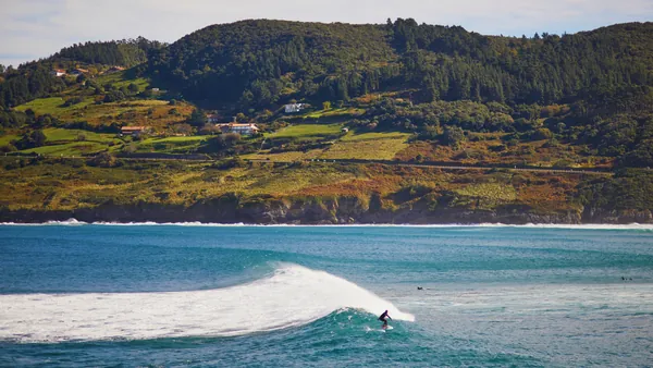 Surfista en la famosa ola de Mundaka con colinas verdes del País Vasco al fondo.