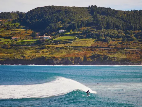 Surfista en la famosa ola de Mundaka con colinas verdes del País Vasco al fondo.