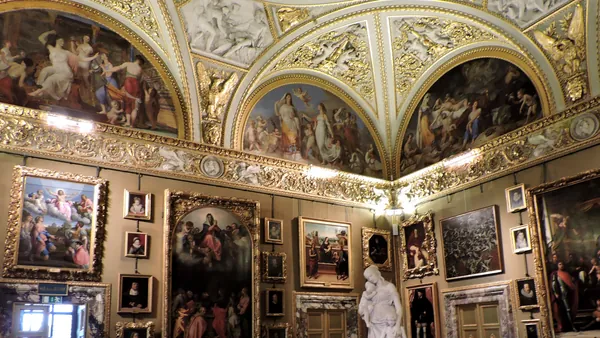 Interior del Palacio Pitti en Florencia, Italia: sala decorada con frescos barrocos en techos abovedados, molduras doradas, numerosas pinturas clásicas en marcos ornamentados y una escultura de mármol blanco.