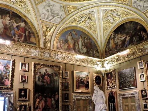 Interior del Palacio Pitti en Florencia, Italia: sala decorada con frescos barrocos en techos abovedados, molduras doradas, numerosas pinturas clásicas en marcos ornamentados y una escultura de mármol blanco.