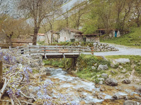 Puente de madera sobre el río Bulnes, en los Picos de Europa, rodeado de casas de piedra y un paisaje natural típico asturiano.