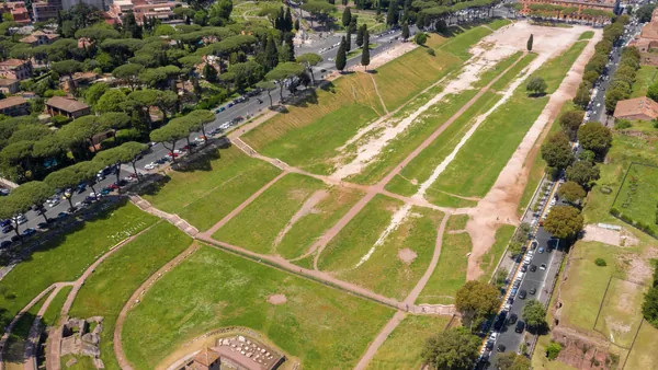 Vista aérea del Circo Máximo en Roma, antiguo hipódromo romano rodeado de árboles y calles, con áreas verdes y restos arqueológicos.