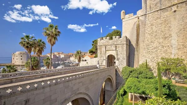 Entrada principal a las murallas de Dubrovnik, con puente de piedra, palmeras y vistas al puerto y a la ciudad amurallada.
