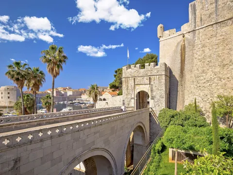 Entrada principal a las murallas de Dubrovnik, con puente de piedra, palmeras y vistas al puerto y a la ciudad amurallada.