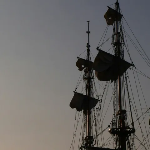 Mástiles y velas de un barco antiguo o réplica al atardecer, siluetas oscuras contra un cielo despejado