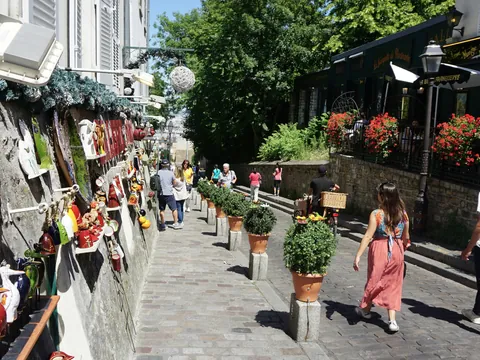 Calle empedrada en Montmartre, París, decorada con macetas y objetos artísticos en las paredes, con personas paseando bajo luz natural y vegetación al fondo.