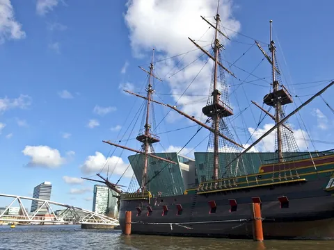Réplica de un histórico barco de vela neerlandés atracado frente al Museo Marítimo de Ámsterdam, con cielo azul y puentes modernos en el fondo.