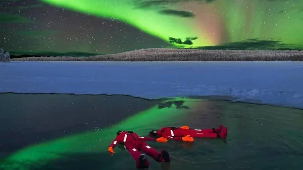 Dos personas con trajes térmicos rojos flotando en agua helada bajo una aurora boreal verde en un paisaje ártico, con nieve y bosque al fondo.