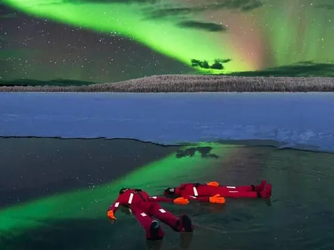 Dos personas con trajes térmicos rojos flotando en agua helada bajo una aurora boreal verde en un paisaje ártico, con nieve y bosque al fondo.