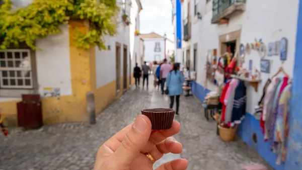 Mano sosteniendo un vaso de chocolate con ginja en una calle típica de Óbidos.
