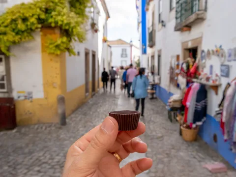 Mano sosteniendo un vaso de chocolate con ginja en una calle típica de Óbidos.