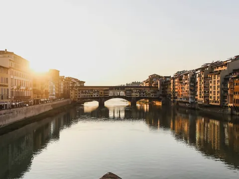 Vista del Ponte Vecchio en Florencia, Italia, al atardecer, con reflejos dorados en el río Arno y edificios históricos a ambos lados.