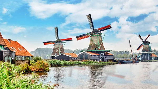 Molinos de viento tradicionales junto a un canal, rodeados de casas con techos rojos y vegetación, bajo un cielo azul con nubes en Zaanse Schans, Países Bajos.