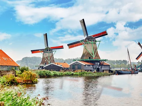Molinos de viento tradicionales junto a un canal, rodeados de casas con techos rojos y vegetación, bajo un cielo azul con nubes en Zaanse Schans, Países Bajos.