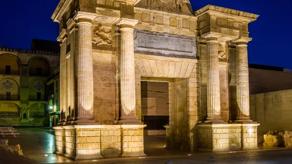Puerta neoclásica iluminada de noche, columnas dóricas, piedra arenisca, cielo azul oscuro, Córdoba