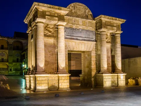 Puerta neoclásica iluminada de noche, columnas dóricas, piedra arenisca, cielo azul oscuro, Córdoba