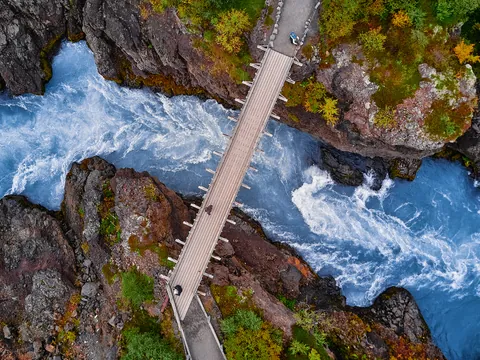 Puente sobre las aguas turquesas del río en Hraunfossar, Islandia, rodeado de rocas volcánicas y vegetación.
